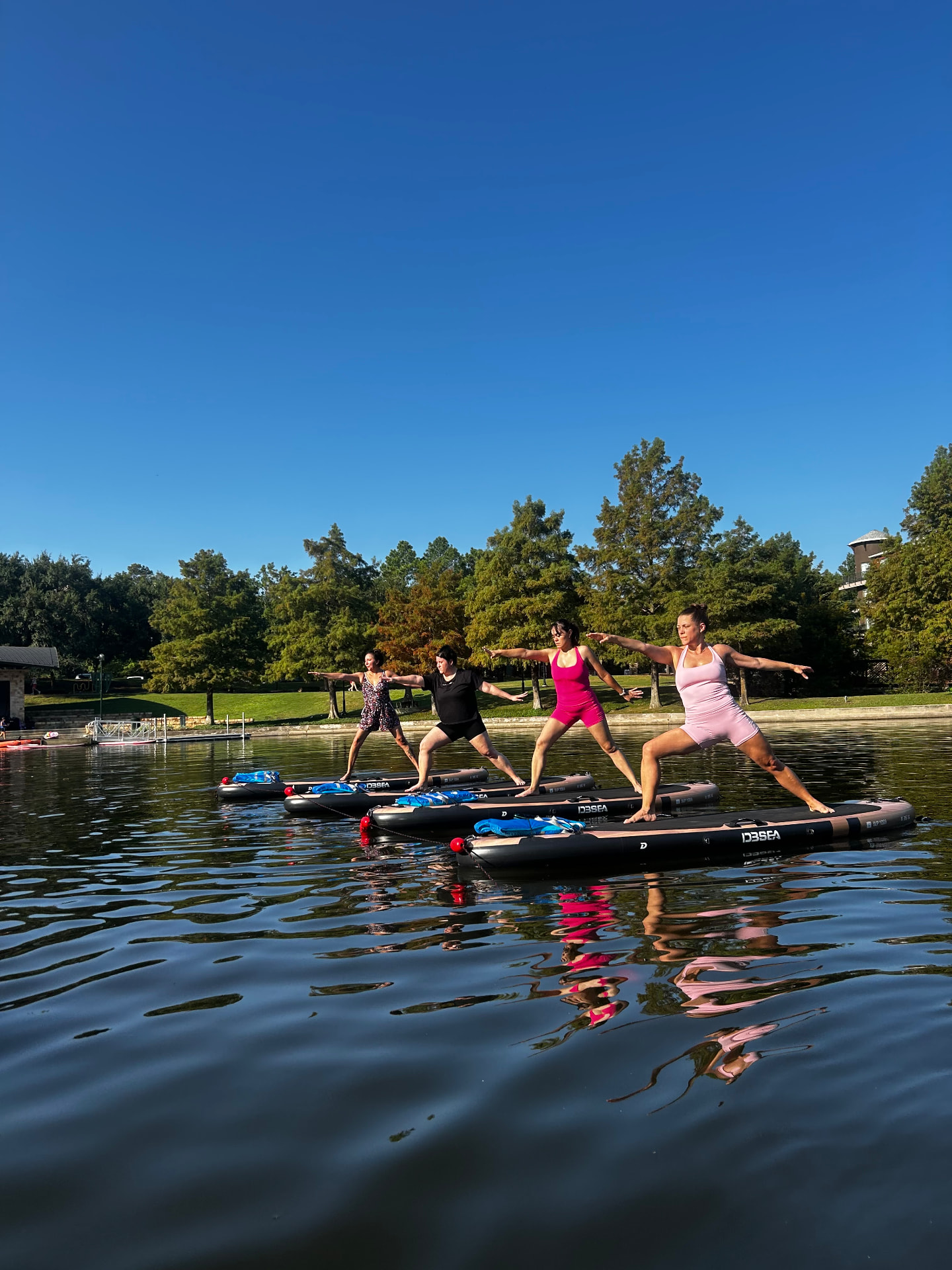 Four women practicing Orbit Flow SUP yoga in warrior pose on paddleboards in a calm lake surrounded by trees