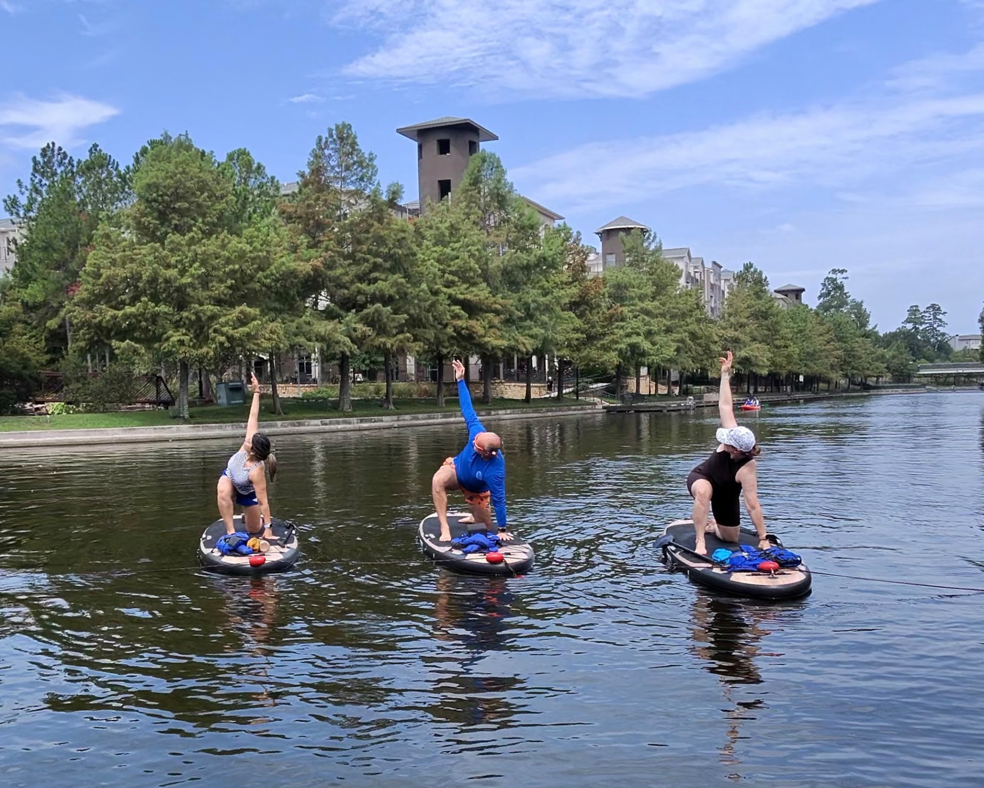 Three people in a low lunge pose with arms extended on paddleboards along a tree-lined waterway