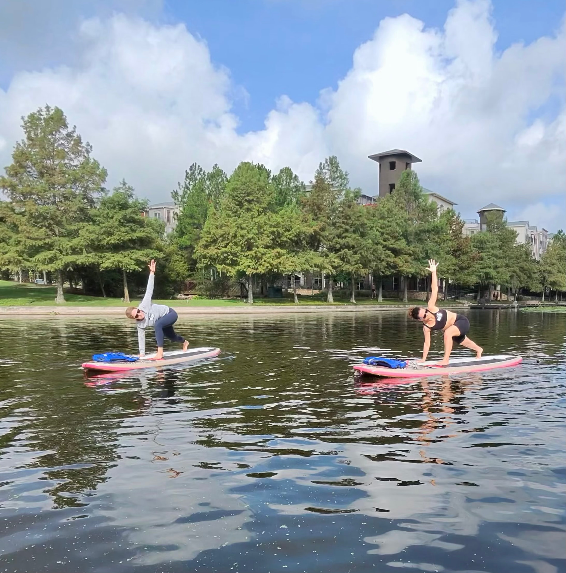 Two women practicing a SUP yoga pose with arms raised on paddleboards in front of a tree-lined lake and tower