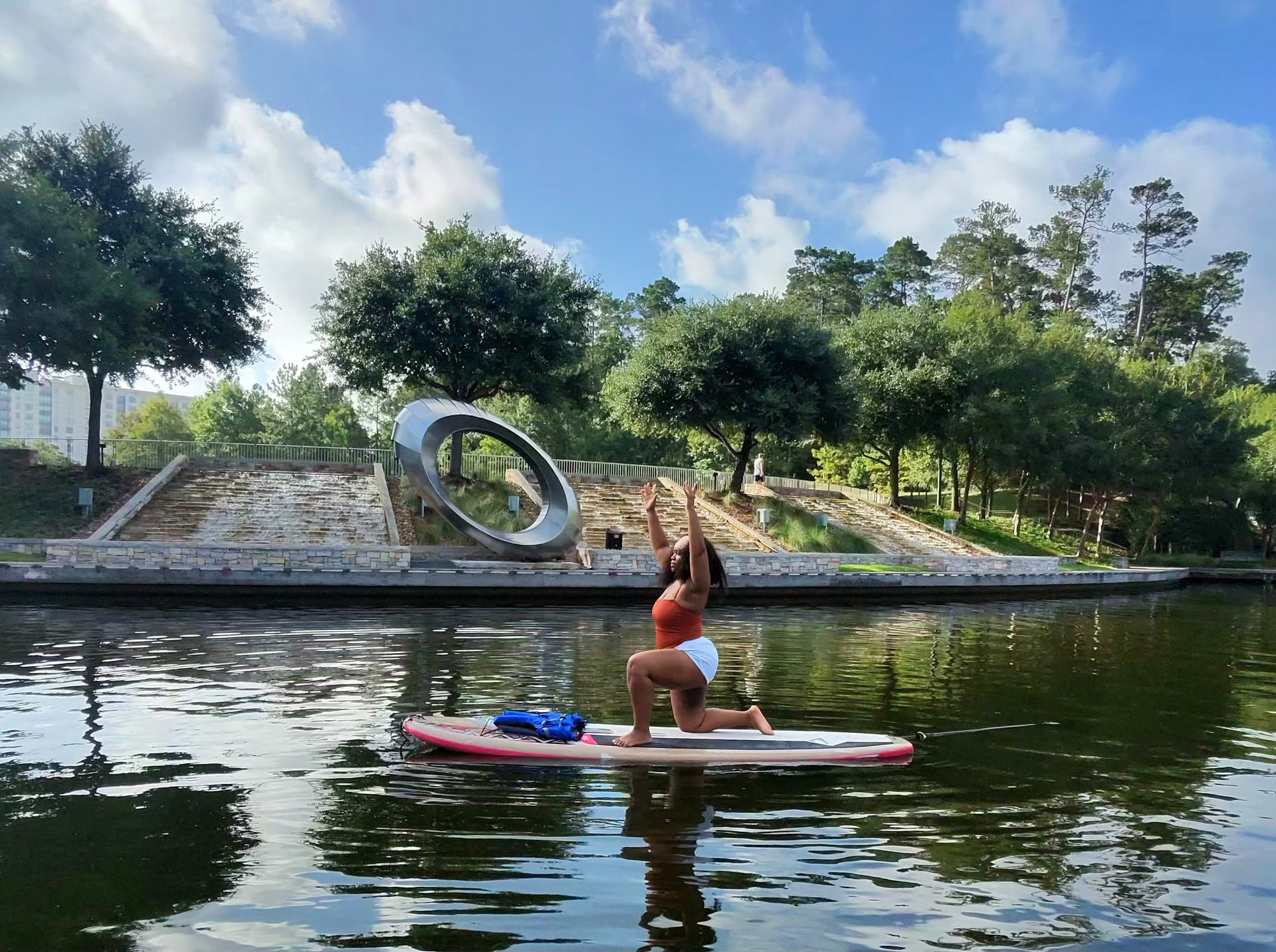 A paddler in crescent lunge with arms raised on a paddleboard in front of the Waterway ring sculpture