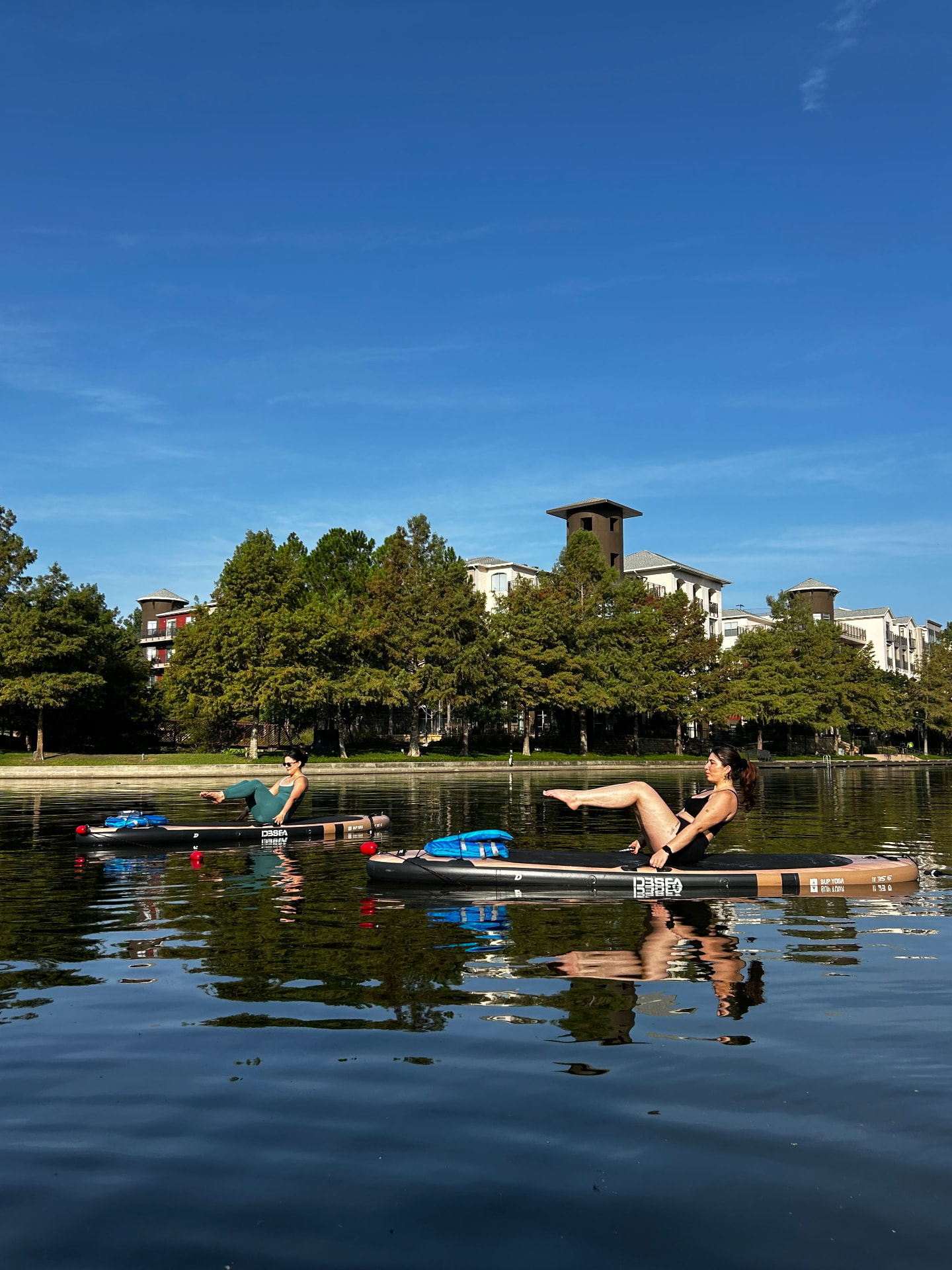 Two women holding a boat pose balance on paddleboards on a calm lake